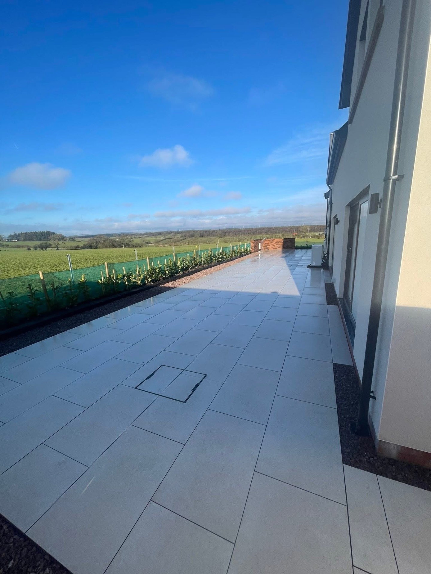 After — long-format grey porcelain terrace with a recessed access cover, looking out over open fields and a new hedging line