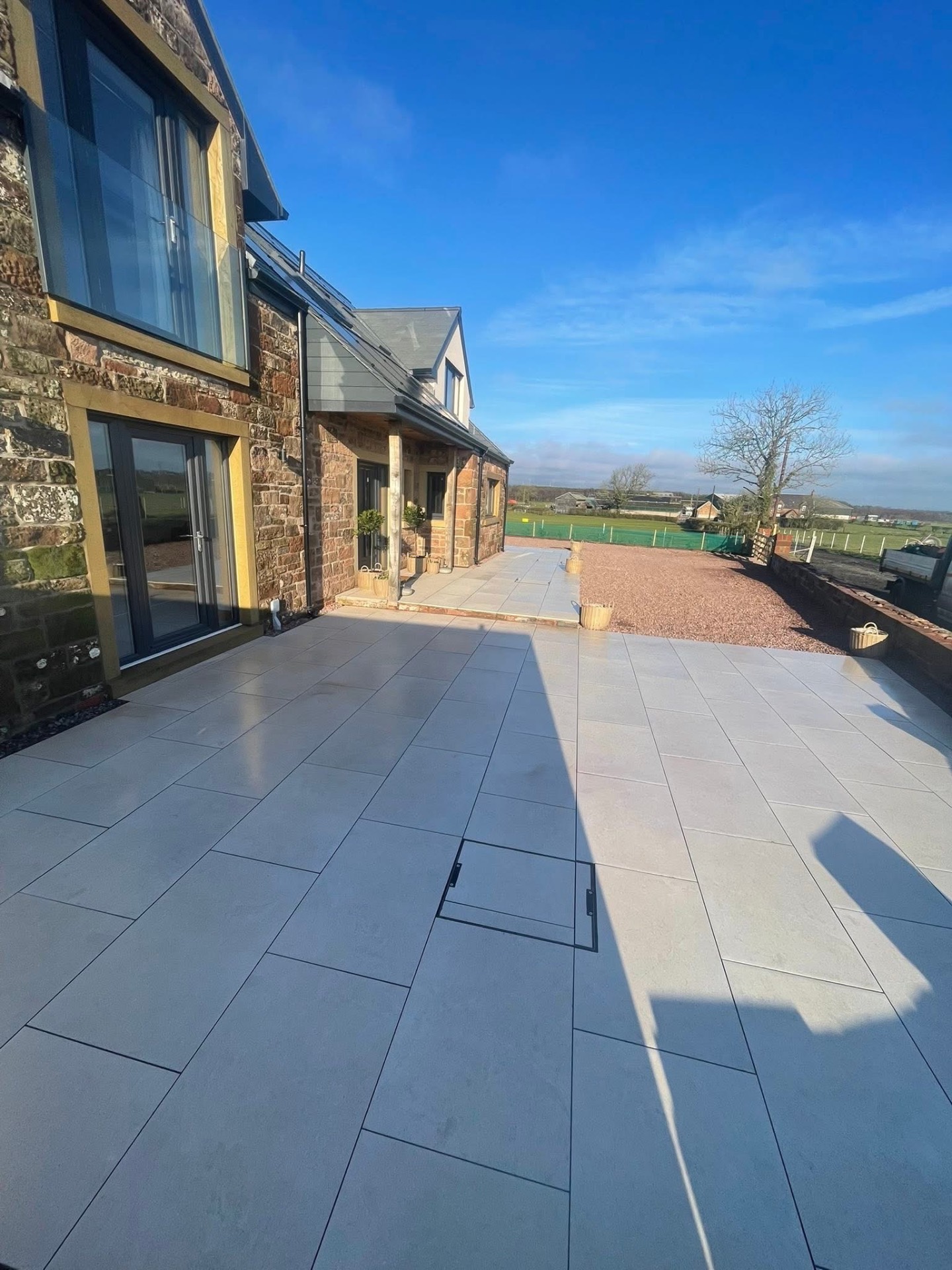 After — side elevation of the stone farmhouse with porcelain terrace continuing past oak-framed bifold doors out toward open fields